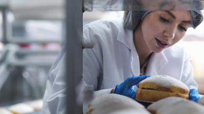 Una mujer con bata y guantes inspecciona panes en una estantería de una panadería o laboratorio.