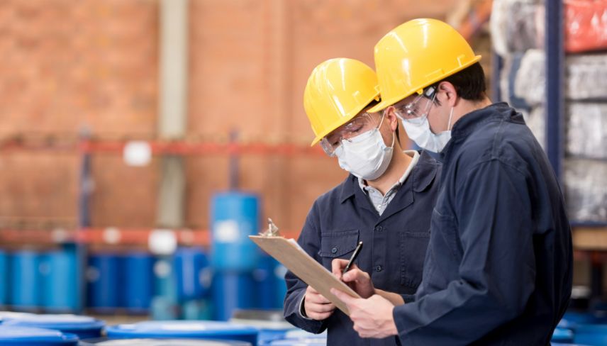Industrial compliance inspection at a chemical manufacturing facility, with two workers in safety helmets and face masks reviewing documents near storage drums.