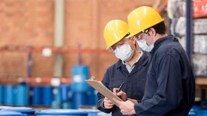 Industrial compliance inspection at a chemical manufacturing facility, with two workers in safety helmets and face masks reviewing documents near storage drums.